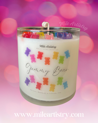 Candle with colorful gummy bear design and 'Mile Artistry' branding on a blurred pink and purple background.