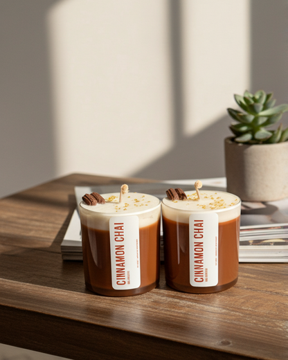 Two candles labeled 'Cinnamon Chai' on a wooden table with a plant in the background.