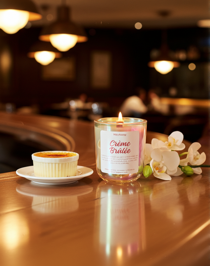 Candle labeled 'Crème Brélée' next to a small dish of crème brélé on a wooden table with a blurred restaurant setting.