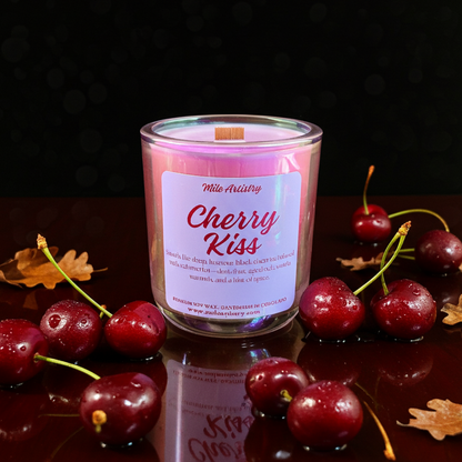 Candle labeled 'Cherry Kiss' surrounded by cherries on a reflective surface.